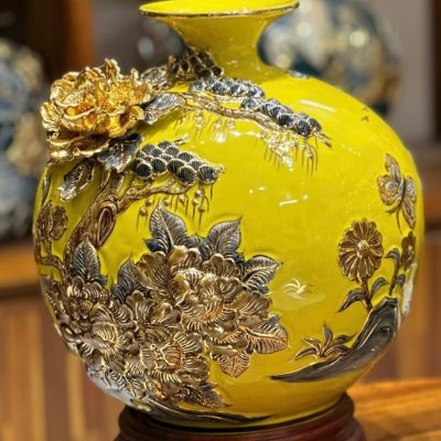 A vibrant yellow Bat Trang ceramic vase featuring an ornate embossed design with 24K gold-painted flowers, pine trees, and a butterfly. The large, round vase with a flared neck sits on a dark wooden base, showcasing intricate Vietnamese craftsmanship.