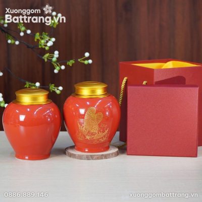 Hộp Đựng Trà Họa Tiết Kim Long Men Đỏ XG-HDT10 bằng gốm sứ Bát Tràng cao cấp, với hai hũ trà màu đỏ rực rỡ nắp vàng kim sang trọng đặt trên bàn gỗ sáng. Một hũ trà có họa tiết cá Rồng (Kim Long) vàng tinh xảo, tượng trưng cho tài lộc. Kèm theo là bộ túi và hộp quà màu đỏ cùng tông, làm nổi bật ý nghĩa phong thủy và giá trị sản phẩm.
