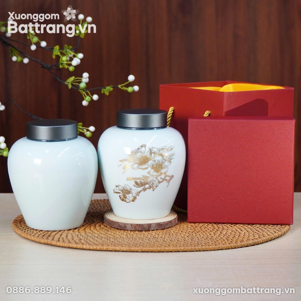 Hai hũ đựng trà gốm sứ Bát Tràng cao cấp với nắp kim loại màu xám bạc, một hũ màu trắng/xanh nhạt trơn và một hũ màu trắng vẽ họa tiết chim hoa vàng kim tinh xảo, đặt trên chiếu mây tre đan cạnh hộp quà màu đỏ. Cành cây trang trí với quả mọng trắng và xanh làm nền.