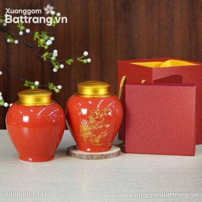 Hai hũ đựng trà gốm sứ men đỏ bóng, nắp vàng đồng, đặt trên mặt bàn gỗ sáng. Một hũ được trang trí họa tiết cây trúc (tre) màu vàng kim, hũ còn lại trơn. Kèm theo là một hộp quà và túi quà màu đỏ với quai vàng ở phía sau, cùng cành cây trang trí quả trắng trên nền gỗ nâu.